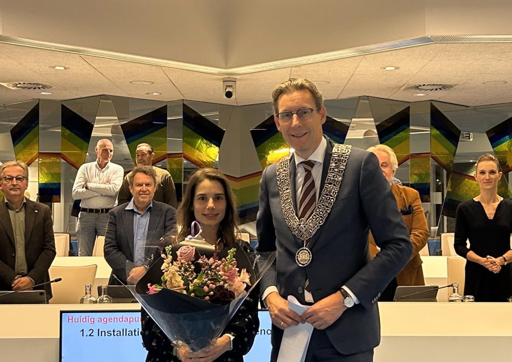 Ima Stokvis staat met een bos bloemen links van Burgemeester Poppens middenin de raadzaal. Op de achtergrond staan diverse raadsleden.