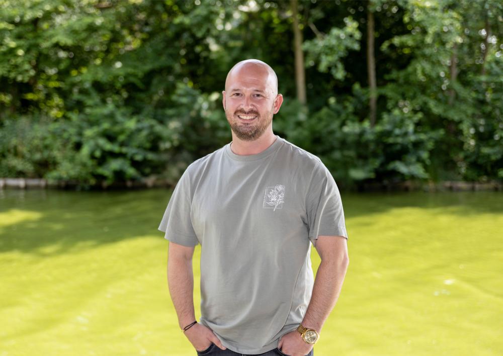 Jongerenwerker Jasper kijkt je lachend aan met op de achtergrond bomen en gras