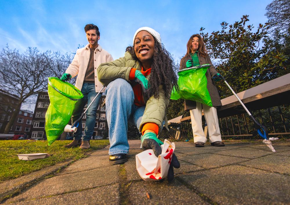 Dame op de voorgrond pakt een prop papier van de grond. Twee mensen op de achtergrond lopen met zakken en grijpers