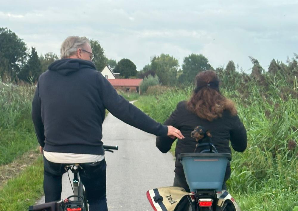 Kees Noomen op de fiets in de polder naast zijn vrouw, van achteren gefotografeerd met een hond achterop de fiets bij zijn vrouw in een mandje. Hij steekt zijn hand uit naar haar
